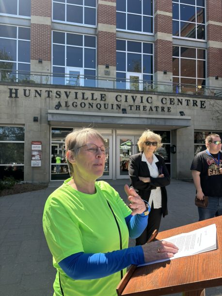 A woman in a bright green T-shirt speaks at a podium in front of a building, the Huntsville Civic Centre