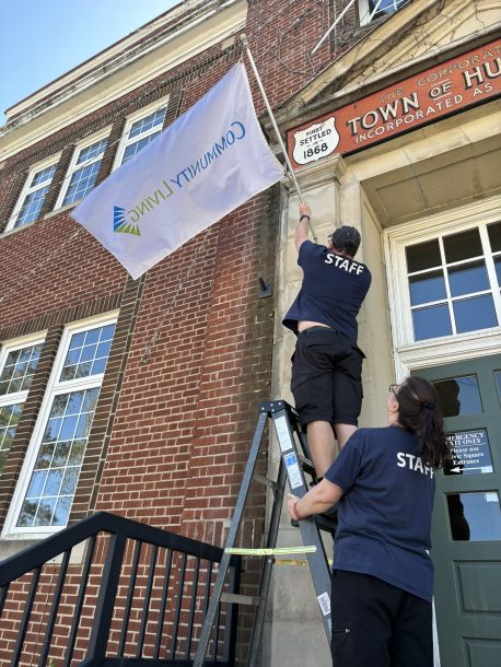 2 people in blue T-shirts that read "staff" across the back, one holds a ladder while the other standing on the ladder and places a flag on a pole into a holder on the side of a building, town hall.