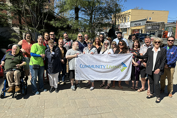 A large group of people standing outside in a community square holding a large, white flag that shows the Community Living logo.