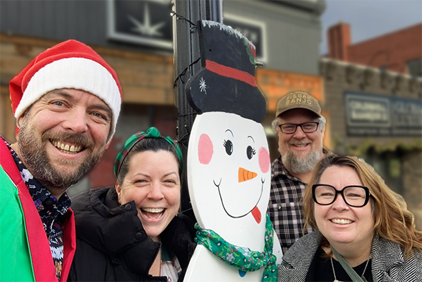 4 smiling adults taking a selfie with a painted wooden snowman on a downtown street.