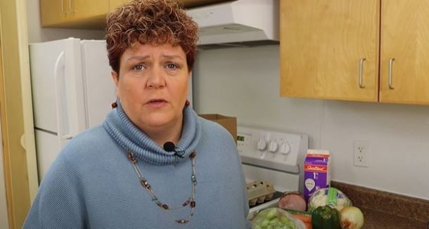 A woman in a blue turtle-neck sweater and a serious facial expression stands in a kitchen. There is a small amount of groceries on the countertop behind her.