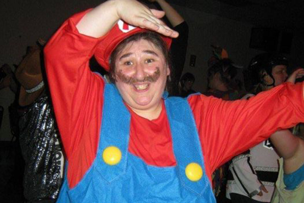 A smiling woman dressed as Super Mario dances in a room filled with other dancing people.