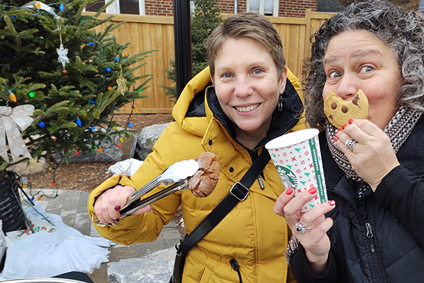 2 smiling woman in a downtown community square. One uses kitchen tongs to hold a cookie while the other holds a paper coffee cup and holds a partially eaten cookie in front of her mouth.