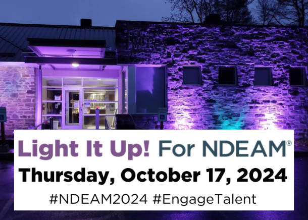 A stone building, Community Living Huntsville's administration offices, lit at night with purple and blue floodlights. Text reads, "Light It Up for NDEAM, Thursday, October 17, 2024".