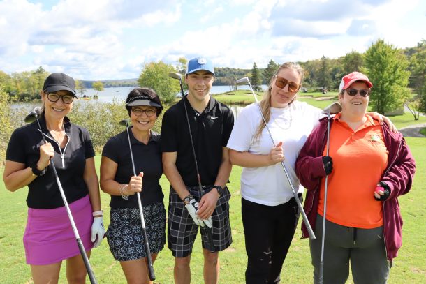 A team of golfers, 4 women and 1 man, pose for a photo with their golf clubs resting on their shoulders while standing at the tee.