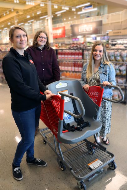 3 women gather around a red shopping cart that has a full-sized grey bucket seat built into it near the handle bar.