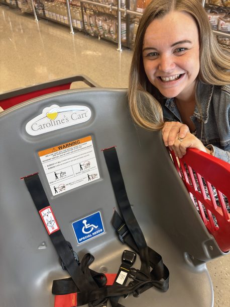 A woman smiles as she crouches beside a shopping cart modified with a full-sized grey bucket seat near the handle bars.