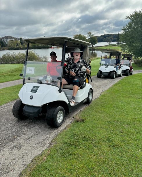 2 men smile as they drive by in a golf cart. 1 man is giving a thumbs up.