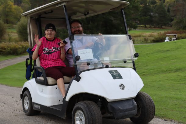 A woman and a man smile and wave while driving by on a golf cart.