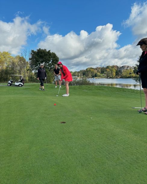 A woman in a hot pink sweater and hot pink hat putts a hot pink ball into a golf green cup.