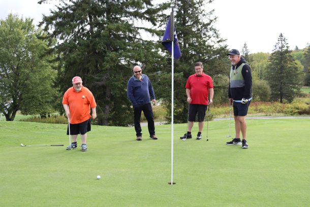 A man in an orange T-shirt finishes a putt while 3 teammates look on in excitement.