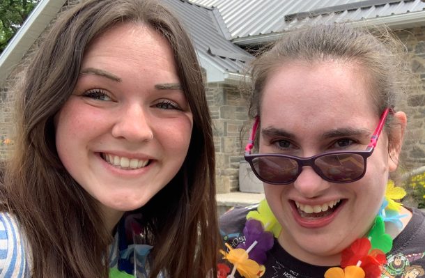 2 smiling woman snap a selfie. The woman on the right is wearing sunglasses and a colourful flower necklace.