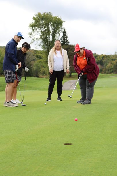 A woman in a maroon sweater putts a pink ball into a golf green cup as 3 teammates watch with excited expressions on their faces.