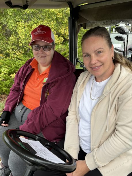 2 smiling women seated in a golf cart. There are golf clubs seen behind them.