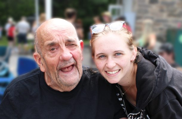 A smiling man in a black T-shirt is seated beside a smiling woman in a black hoodie. They are at an outdoor party.