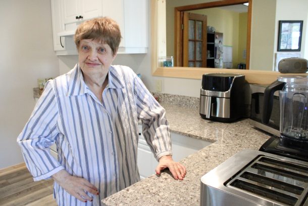 A smiling woman rests one hand on a brightly speckled countertop and another on her hip while standing in a bright kitchen with white cabinets.