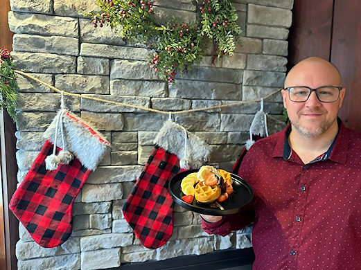 A smiling man in a red button-up shirt and glasses holds a plate of Belgian waffles and fresh fruit, while standing in front of a stone fireplace decorated with an evergreen wreath and 2 red-plaid stockings.