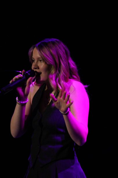 A young woman with blonde hair stands under a purple spotlight and sings into a microphone.